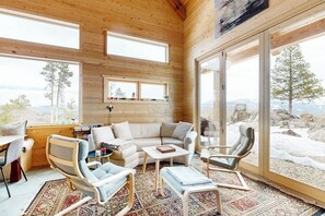 A cozy cabin interior with a wooden interior, a sofa, chairs, a small table, and large windows offering a view of the outdoors. - Modern off grid cabin with stocked kitchen & private mountain view- dog friendly (Tabernash)