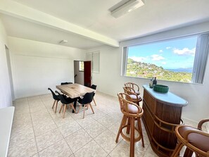 A dining area with a wooden table and chairs, a bar area with a counter, and a large window with a view of greenery and hills.