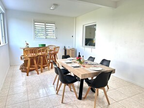 A dining area with a wooden table set for six, surrounded by black chairs, a wicker chair set, and a window with blinds. - Appartement Cannelle - Villa Mathéo - Les Trois Ilets 5 min from the beach (Les Trois-Îlets)