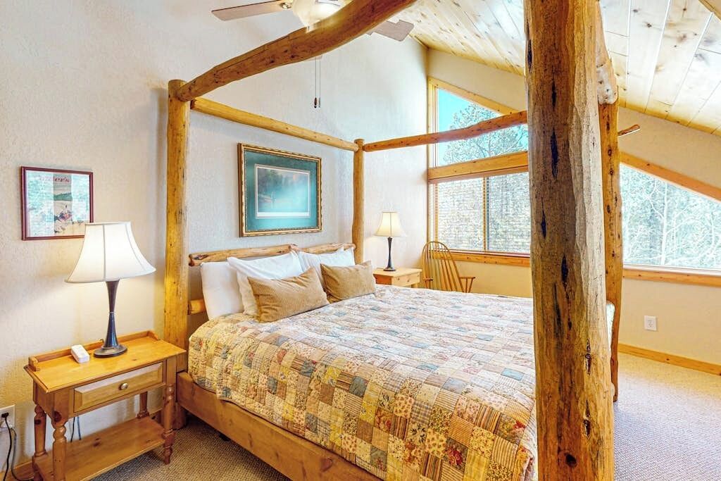 Een slaapkamer met een houten hemelbed, twee bedlampjes, een nachtkastje en een raam met uitzicht op bomen.