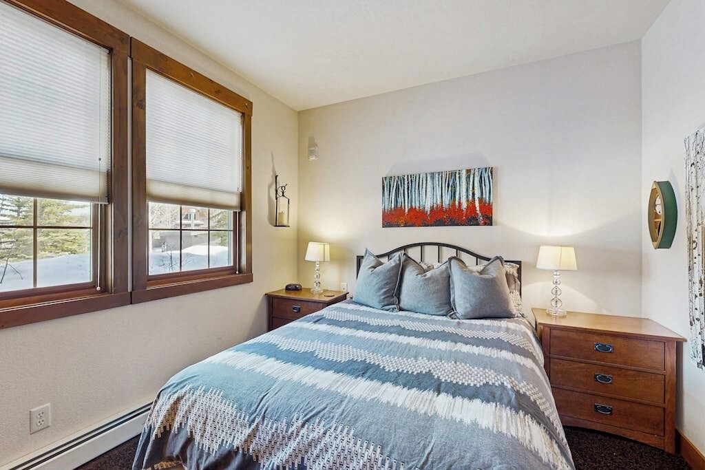 Une chambre à coucher comprenant un lit, deux tables de chevet, une œuvre d’art accrochée au mur et une fenêtre avec des stores.