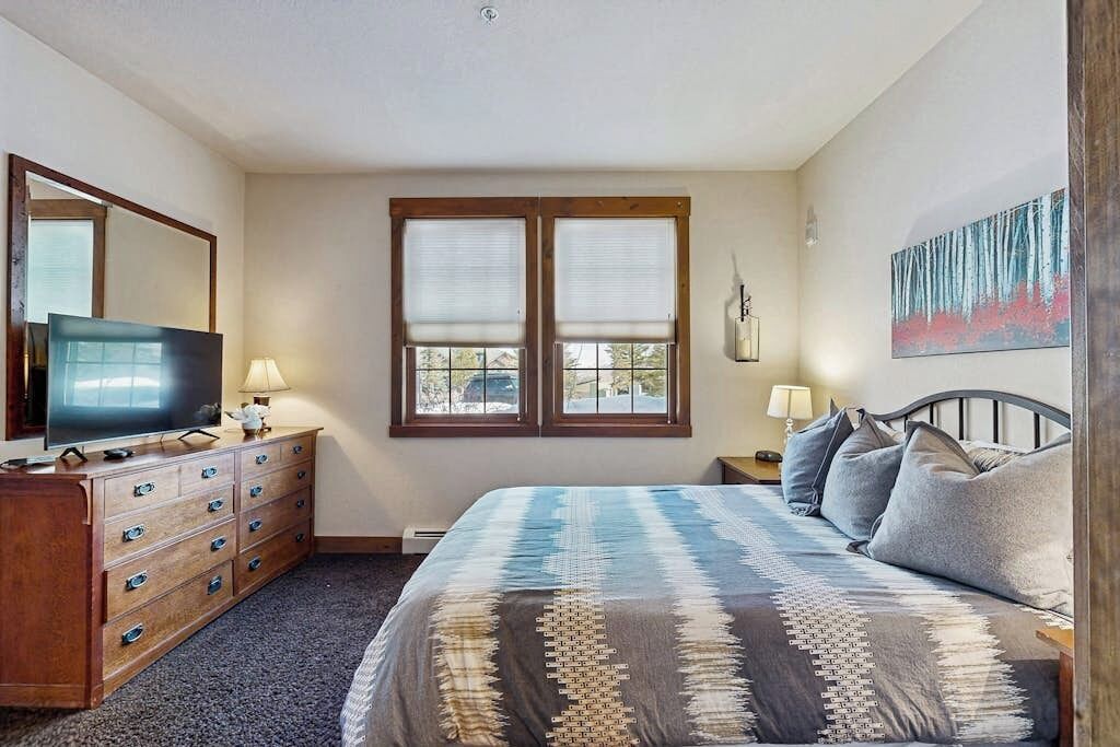 Un dormitorio con cama, cómoda, televisor, dos ventanas con persianas, un cuadro en la pared y una lámpara en la mesita de noche.