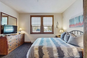Ein Schlafzimmer mit einem Bett, einem Schminktisch, einem Fernseher, zwei Fenstern mit Jalousien, einem Bild an der Wand und einer Lampe auf dem Nachttisch.