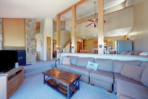 A living room with a sofa, coffee table, TV, stone wall, wooden beams, and a kitchen in the background.