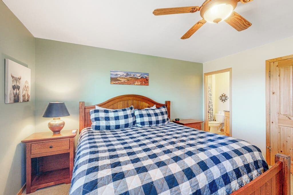 Une chambre à coucher avec un lit en bois, une table de chevet avec une lampe, un ventilateur de plafond et une porte donnant sur la salle de bain.