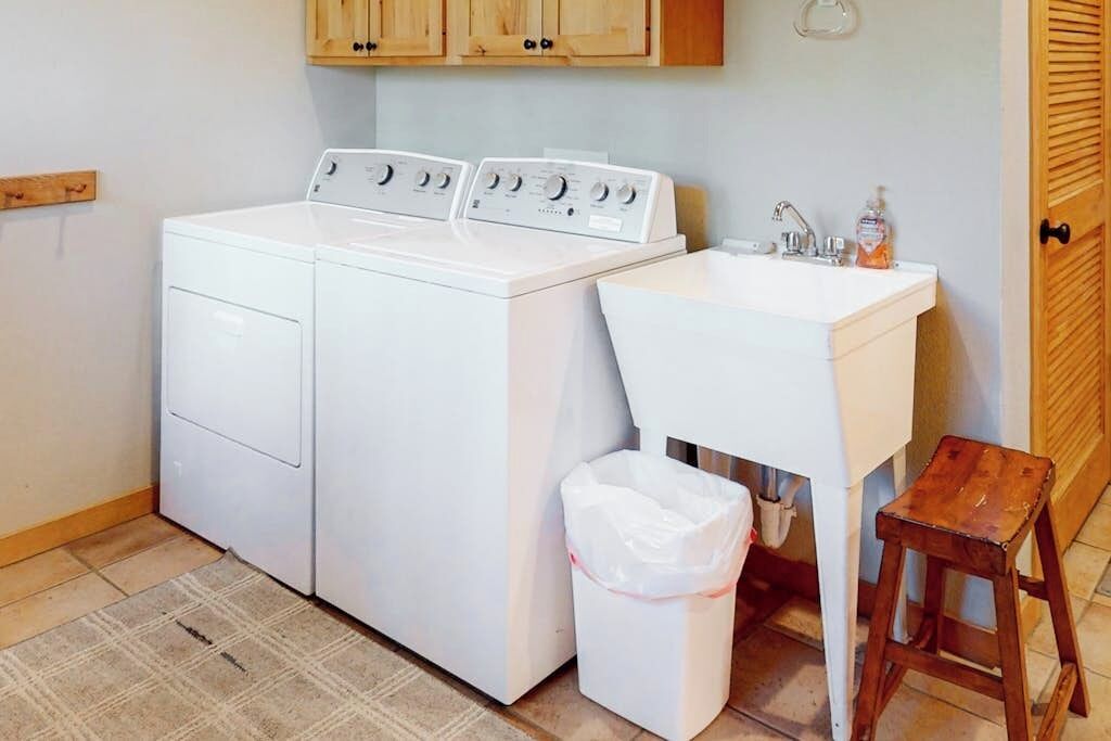 Un espace buanderie comprenant un lave-linge, un évier, un tabouret en bois et une poubelle.