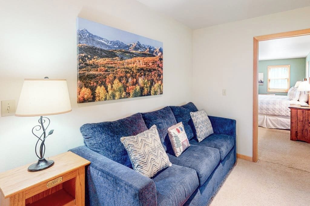 A living room with a sofa, pillows, a lamp, and a painting of a mountainous landscape.