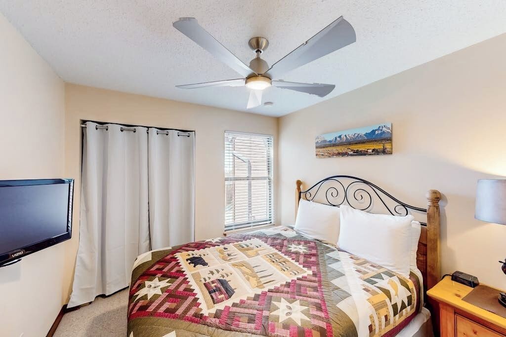 A bedroom with a bed, a ceiling fan, a window with blinds, a painting on the wall, and a television on the wall.