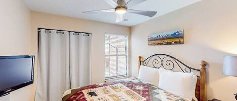 A bedroom with a bed, a ceiling fan, a window with blinds, a painting on the wall, and a television on the wall.