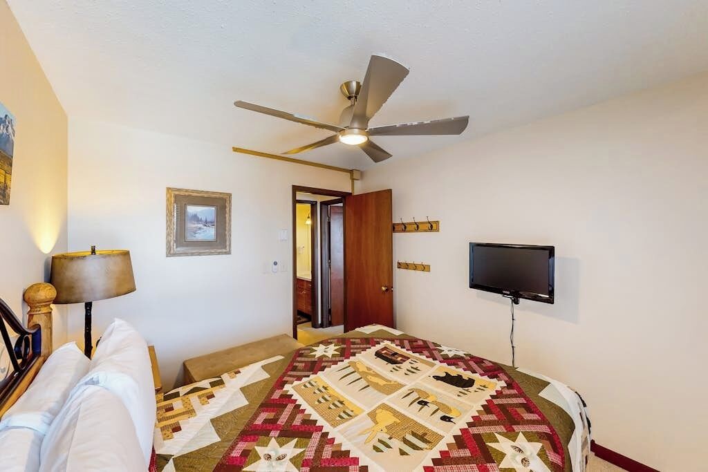 A bedroom with a bed, a ceiling fan, a television, and a door leading to another room.