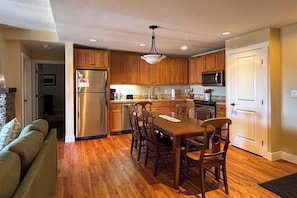 A modern kitchen with wooden cabinets, a stainless steel refrigerator, a dining table with chairs, and a microwave. - 310 Base Camp | Condo in Granby (Granby)