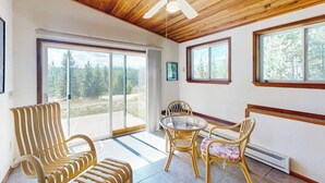 Ein Zimmer mit Holzboden, Deckenventilator, Glastisch mit Stühlen und großen Fenstern mit Blick auf Bäume.