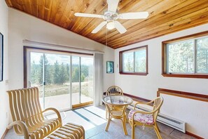 Une pièce avec un plafond en bois, un ventilateur de plafond, une table en verre avec des chaises et de grandes fenêtres donnant sur la forêt.