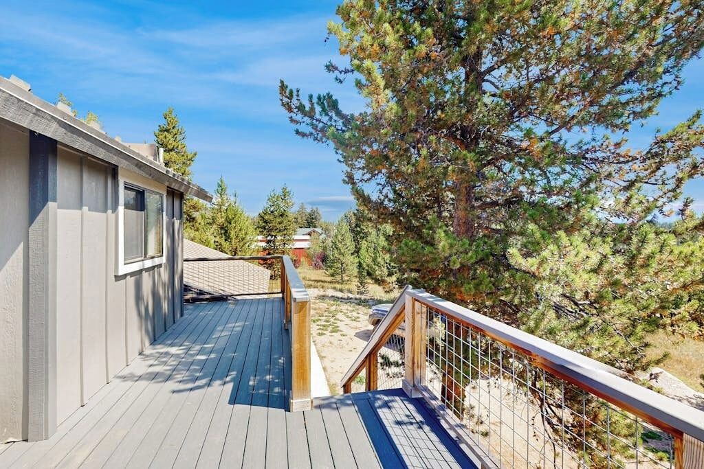 Una terraza de madera con barandilla, un edificio con ventana y un árbol grande al fondo.