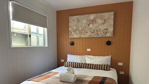 Un lit bien fait, avec un oreiller à rayures et une serviette enroulée, une œuvre d’art accrochée au mur et une fenêtre avec des stores.