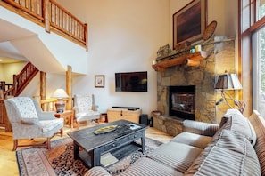 A living room with a stone fireplace, a television mounted on the wall, and a staircase leading to an upper level. - Walk-to-Lift Townhome w/Fireplace, W/D, Shared Hot Tubs, Pool, and Firepit (Steamboat Springs)