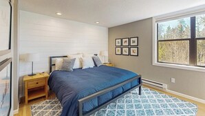 Un dormitorio con cama, mesitas de noche, una ventana y cuadros en la pared.