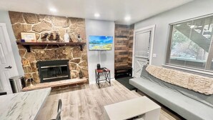 Ein modernes Wohnzimmer mit Kamin aus Stein, einem Wand-Fernseher, einem grauen Sofa und Holzboden.