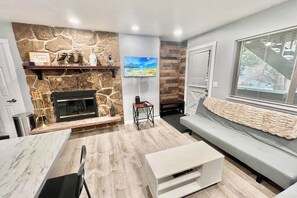 Ein modernes Wohnzimmer mit Kamin aus Stein, einem Wand-Fernseher, einem grauen Sofa und Holzboden.