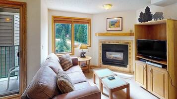 A living room with a sofa, a fireplace, a television, and a view of trees through large windows.