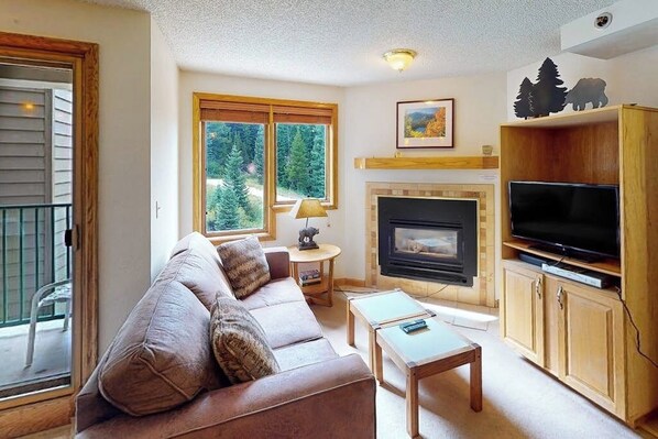 Ein Wohnzimmer mit einer Couch, einem Kamin, einem Fernseher und Blick auf Bäume durch große Fenster.