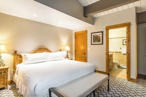 Quarto com cama grande, luminárias de cabeceira, porta de madeira e banheiro visível por uma porta aberta.