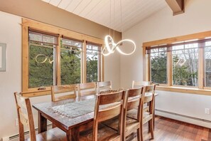 Uma área de refeições com mesa e cadeiras de madeira, janelas amplas com vista para árvores e um lustre.