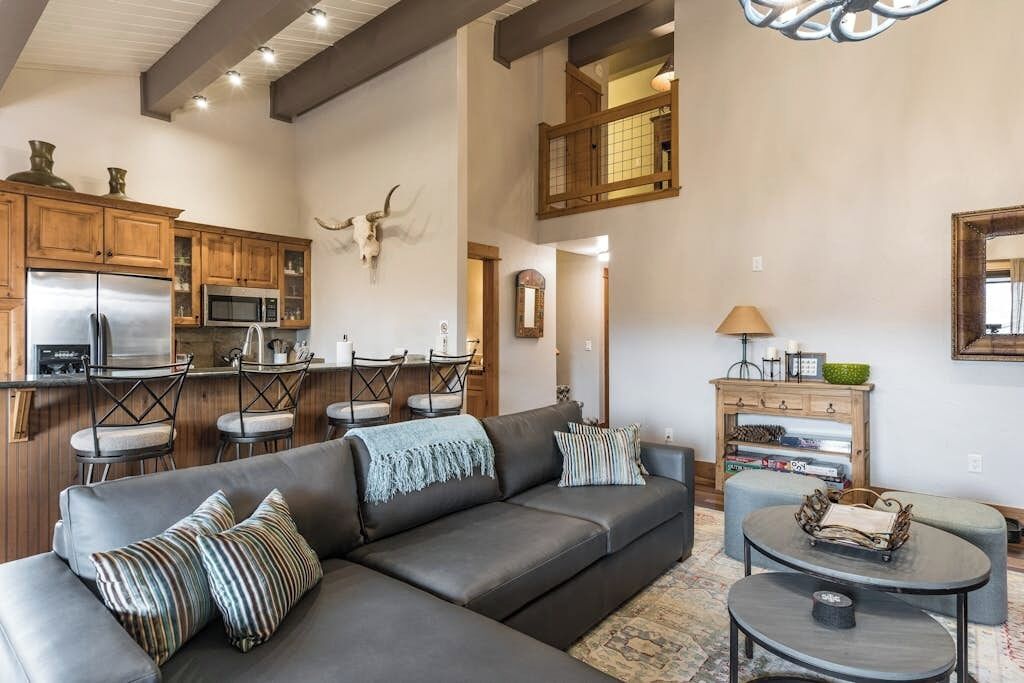 A modern living room with a sectional sofa, a dining area with chairs, a kitchen with appliances, and a loft space with a view.