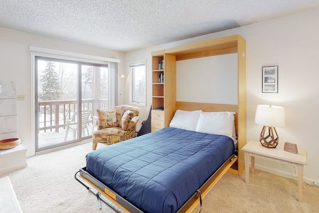 Une chambre à coucher comprenant un lit, une chaise, une table de chevet, une lampe et une porte-fenêtre coulissante donnant sur un balcon.