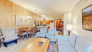 Een woonkamer met een houten wand, een eethoek met stoelen en een tafel, een bank, een salontafel en een keuken op de achtergrond.