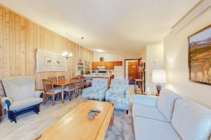 Ein Wohnzimmer mit einer Holzwand, einem Essbereich mit Stühlen und einem Tisch, einer Couch, einem Couchtisch und einer Küche im Hintergrund.