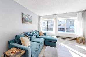 Ein modernes Wohnzimmer mit einem Ecksofa, einem kleinen Holztisch, einer Wanddekoration und einem großen Fenster mit Vorhängen.