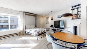 Quarto de hotel moderno com cama, escrivaninha, cadeira, televisão e janela com vista para prédios.