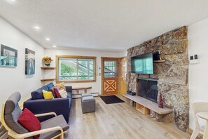 Ein Wohnzimmer mit Kamin aus Stein, einer Couch, einem Fernseher und einem Fenster mit Blick auf Bäume.