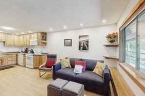 Eine moderne Küche mit Holzausstattung, einer Mikrowelle und einem Herd. Im Wohnbereich befindet sich eine dunkelgraue Couch mit Kissen und ein kleiner Hocker. Ein gerahmtes Bild hängt an der Wand, und ein Fenster bietet Blick ins Freie.