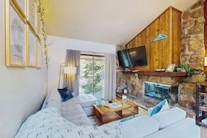Ein Wohnzimmer mit Kamin aus Stein, einem Flachbildfernseher und einem großen Fenster mit Vorhängen.