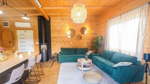 L’intérieur d’un chalet confortable, avec un plafond en bois, un canapé vert, un coin repas avec des chaises et une grande fenêtre agrémentée de rideaux.