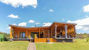 Un bâtiment moderne en bois, doté d’un espace extérieur couvert, d’une grande fenêtre et d’une pelouse verdoyante.
