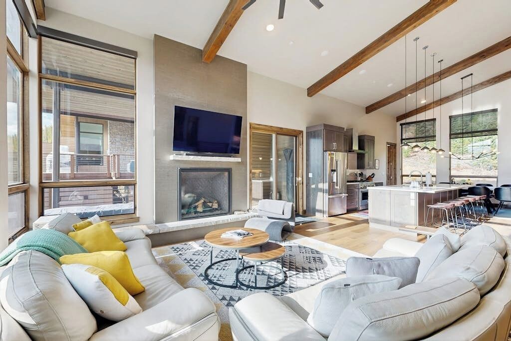 A modern living room with a fireplace, a kitchen area, and large windows.