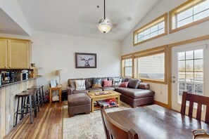 A spacious living area with a sofa, coffee table, and a dining table. There is a kitchen area with bar stools and a ceiling light. - Sunny & Spacious Condo in Downtown Dillon, Fireplace, WiFi (Dillon)