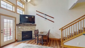 Una sala de estar con chimenea, televisor montado encima, mesa de comedor con sillas y una escalera.