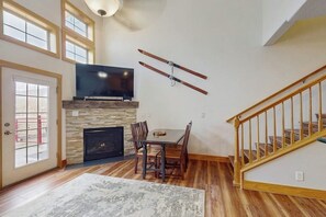 A living room with a fireplace, a TV mounted above it, a dining table with chairs, and a staircase. - Sunny & Spacious Condo in Downtown Dillon, Fireplace, WiFi (Dillon)