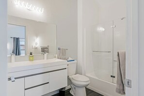 Un bagno moderno con lavandino, specchio e WC bianchi, e una cabina doccia in vetro.