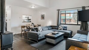 A modern living room with a sectional sofa, a coffee table, and a TV. There is a kitchen in the background. - Two-Level Condo w/ Mountain View, Wood Stove, WiFi, Laundry & Central Location (Pagosa Springs)