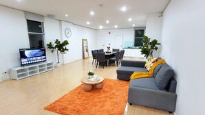 A modern living room with a grey sofa, orange rug, TV, dining table, chairs, and potted plants.