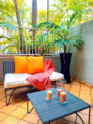 Eine Terrassenfläche mit einem Korbstuhl-Tisch, einem wei ßen Sofa mit orange- und roten Kissen sowie einem Blumentopf.