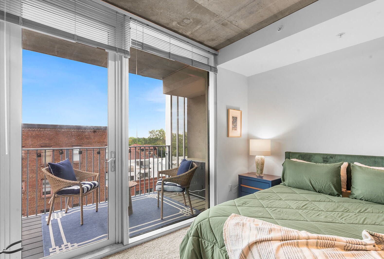 Une chambre d’hôtel avec un balcon, un lit avec une literie verte, une table de chevet avec une lampe et des chaises en osier.