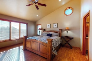 Ein Schlafzimmer mit einem hölzernen Bett, einem Deckenventilator, einem Fenster mit Jalousien, einer Wandlampe und einem kleinen Tisch.