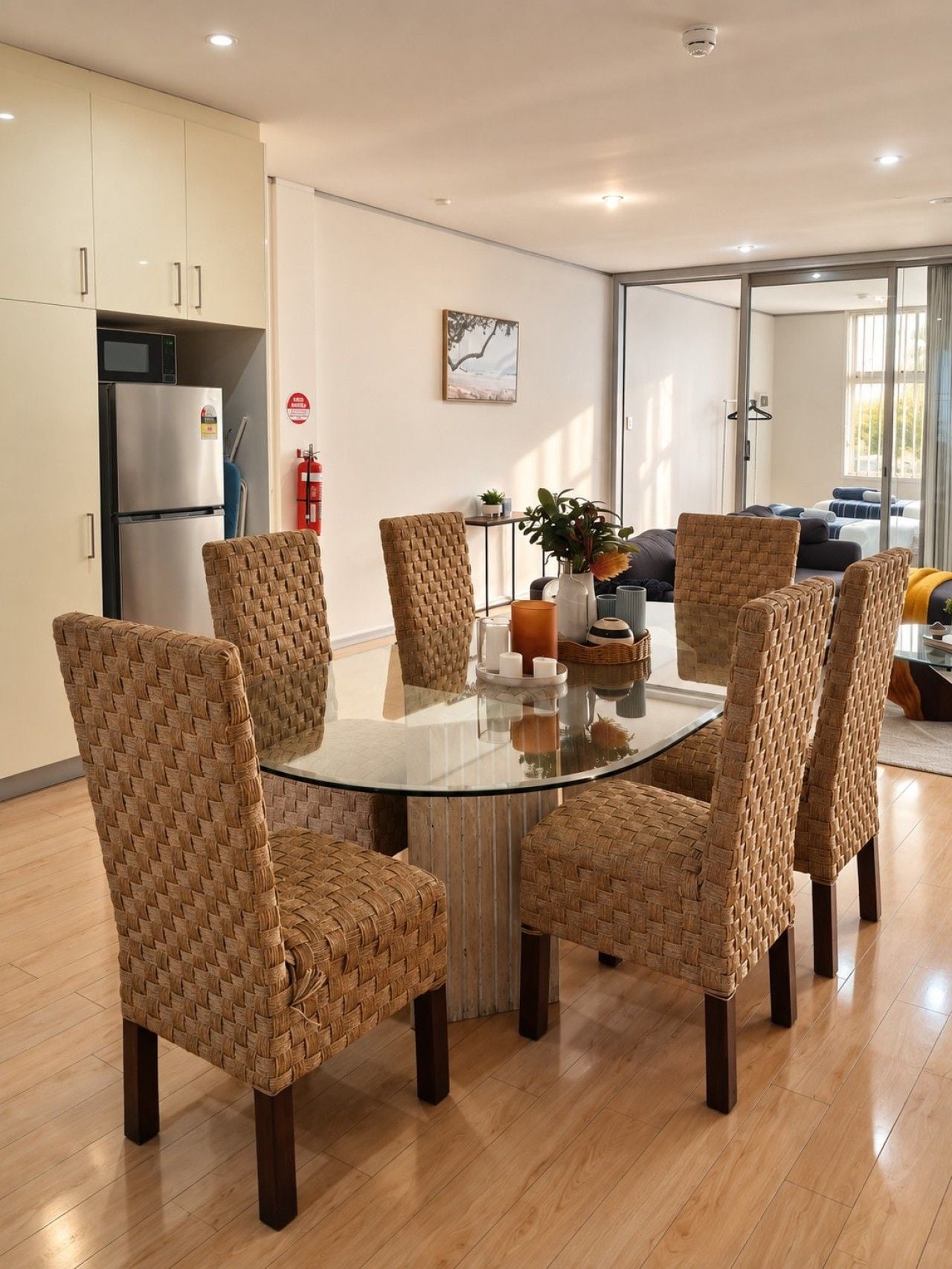 A modern kitchen with a round glass table surrounded by woven chairs, a refrigerator, and a fire extinguisher.