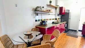 Ein Essbereich mit einem runden Glastisch und Korbstühlen, eine Küche mit Kühlschrank und roten Schränken sowie ein Holzboden.